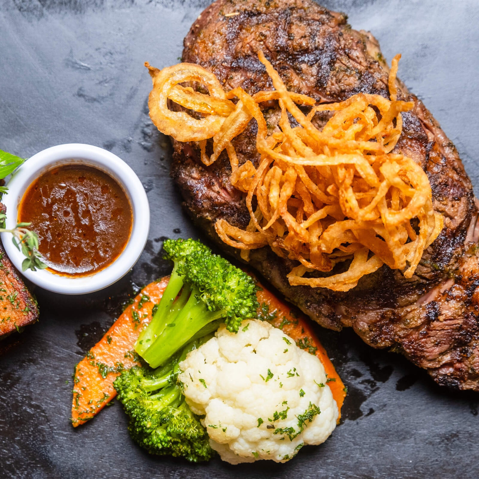 Grilled steak topped with crispy fried onions, served with broccoli, cauliflower, carrots, and a side of sauce.
