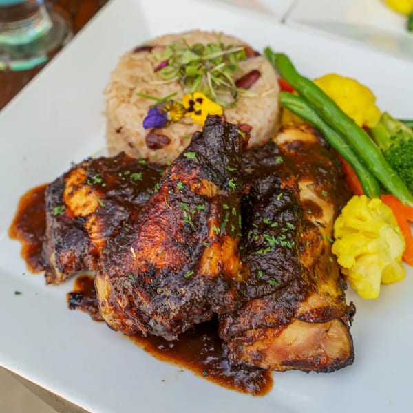A plate of Caribbean jerk chicken with rice and peas, garnished with edible flowers and mixed vegetables.