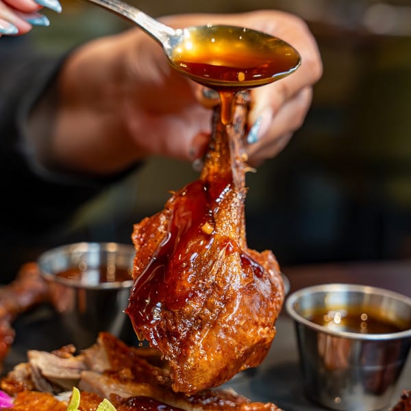 A hand drizzles sweet sauce from a spoon onto a piece of Caribbean jerk chicken.