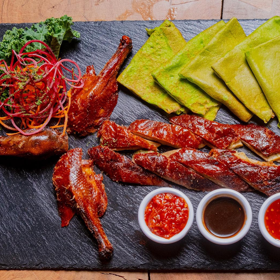Roasted duck, whole and sliced, with green pancakes, kale, and three dipping sauces on a dark platter.