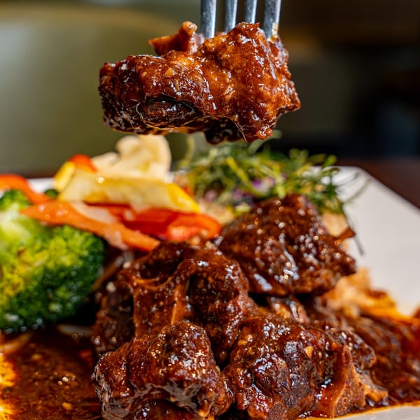 A fork lifts a piece of saucy oxtail from a plate with more oxtail and vegetables.